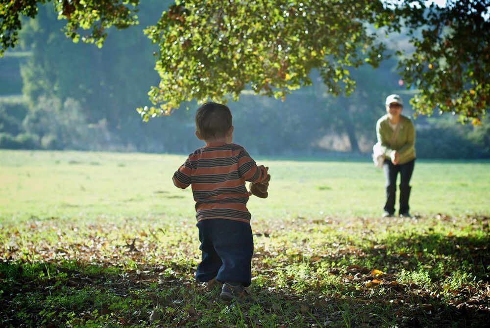 baby walking towards mum in the park who is encouraging her child to come towards her