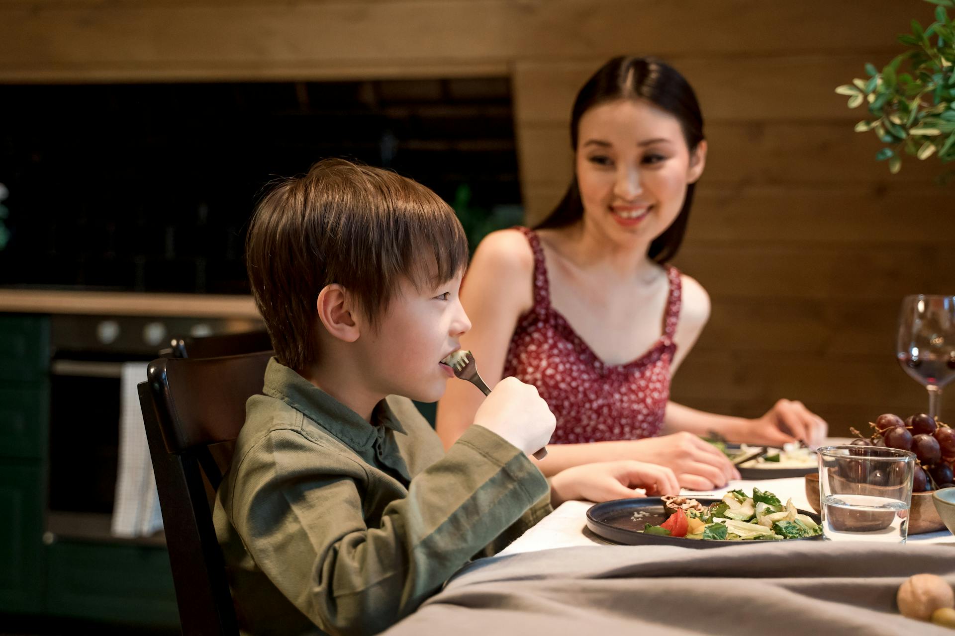 A child eats their dinner at home with their parent smiling at them.