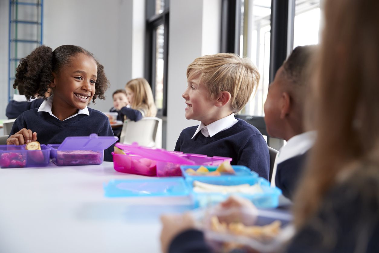 Kids sat around school table eating their lunches from their packed lunch.