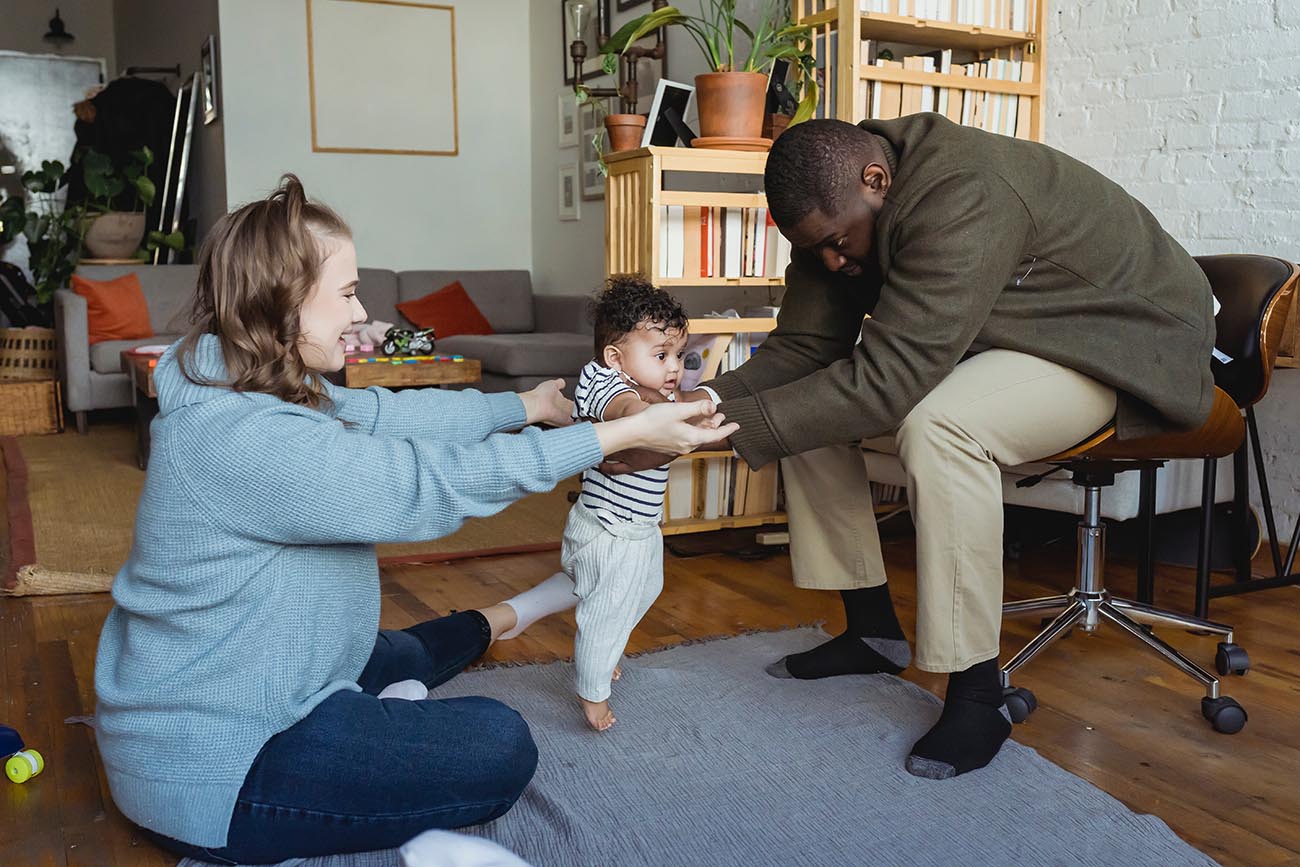 mum and dad helping baby to walk between them both in the lounge