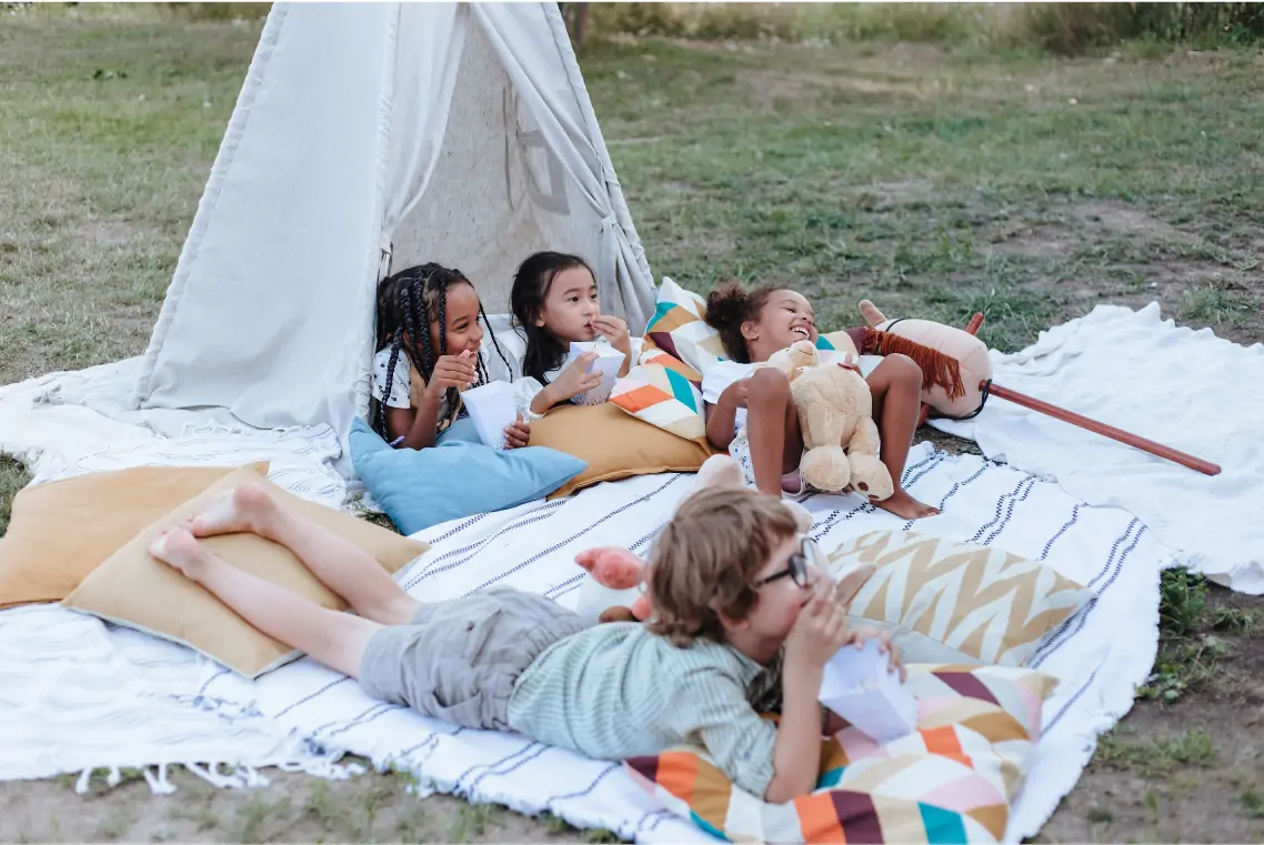 Children sit on cushions and blanket under a garden DIY den.